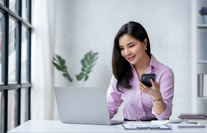 Mulher em escritório utilizando notebook e com celular na mão, sorrindo para a tela e satisfeita com seu serviço de BPO Folha de Pagamento.